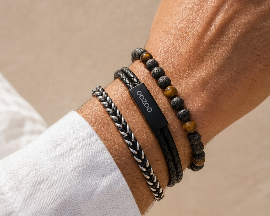 A close-up of a man's wrist wearing four distinct bracelets: a black braided cord bracelet, a black beaded bracelet with brown tiger's eye beads, a black cord bracelet with a black rectangular plate engraved with 'OOOO', and a silver-and-black braided cord bracelet. He is wearing a white collared shirt against a neutral beige background.